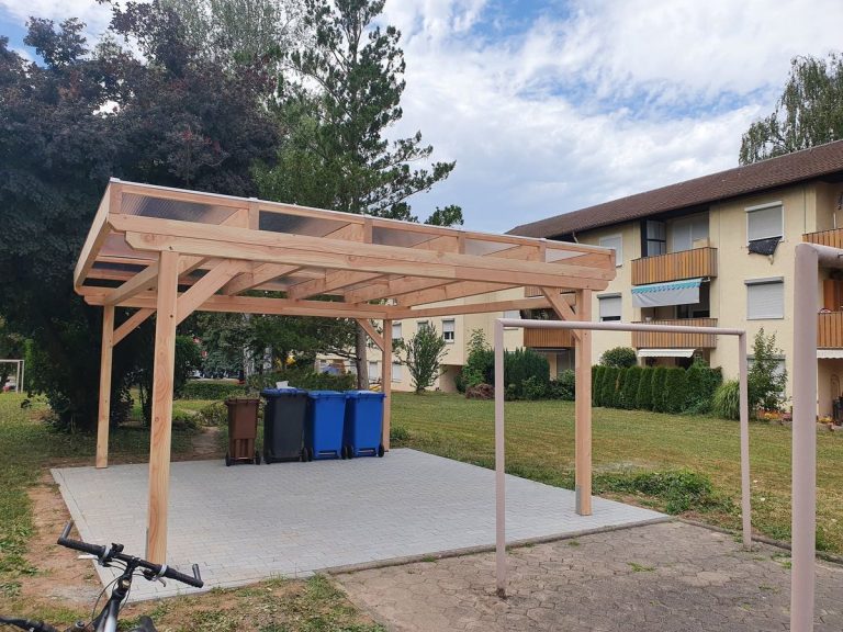 Freistehender Carport aus Holz mit Satteldach auf einer Kiesfläche, im Hintergrund ein Wohnhaus