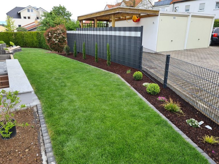 Angelegte Rasenfläche mit Staudenbeet und Rindenmulch entlang eines Sichtschutzzauns